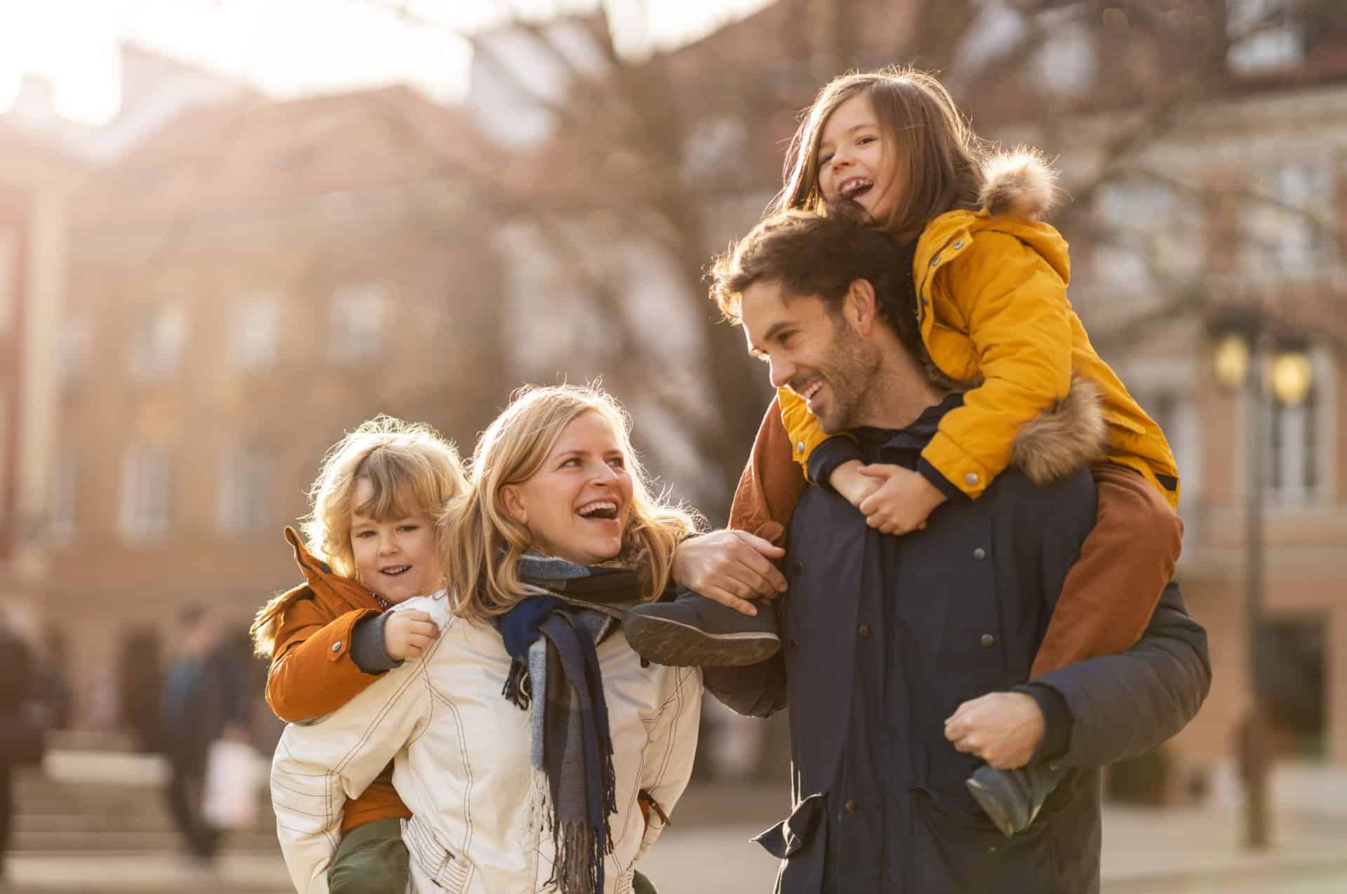 A husband and wife both give piggybacks to their young children in the middle of a city.